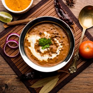 A close-up of creamy dal makhani served with fresh cream and garnished with cilantro, surrounded by spices and onion slices on a wooden surface.